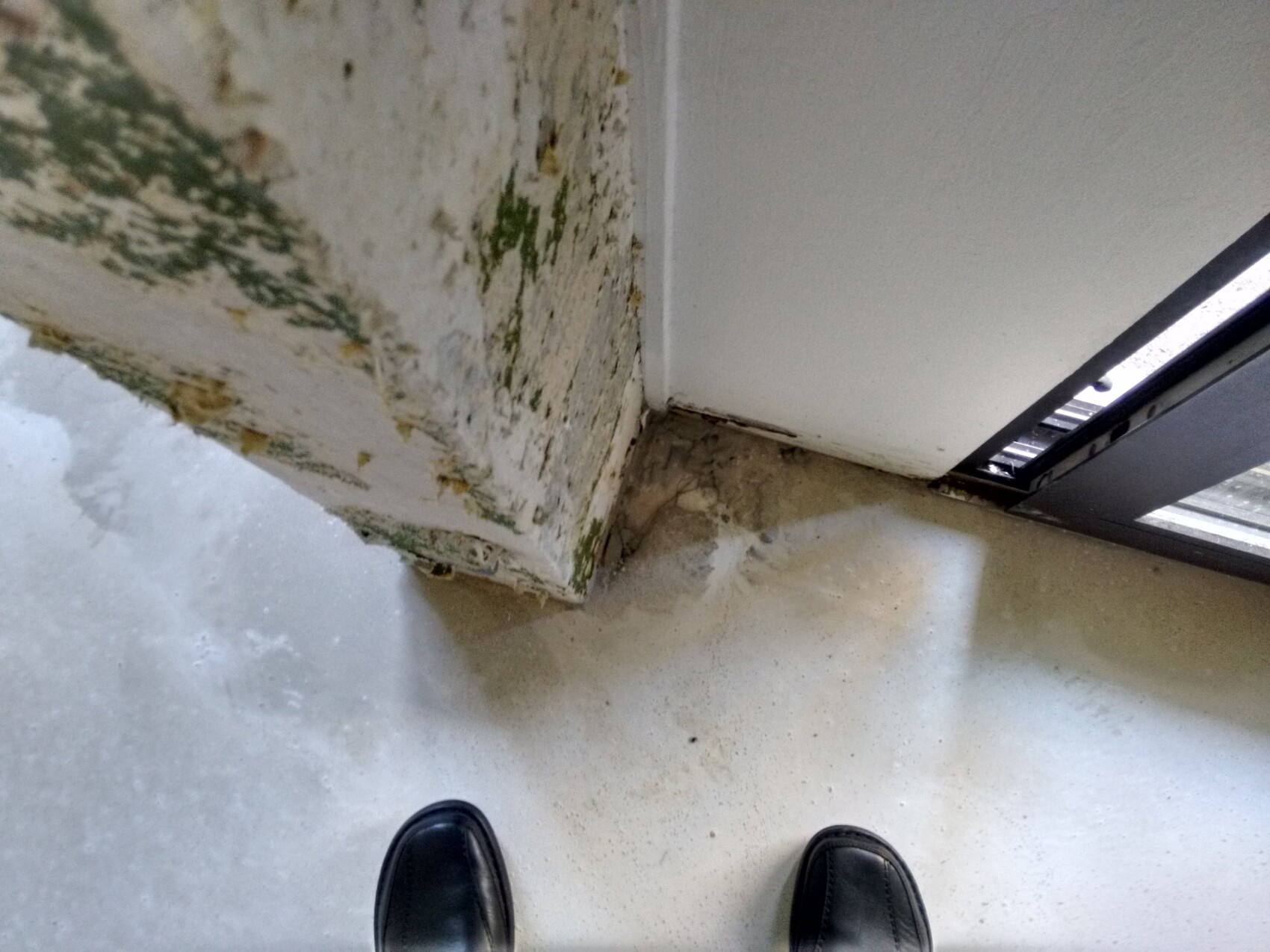 Feuchte Mauer mit Schimmel am Türrahmen, Bodenplatte und Schuhe im Vordergrund