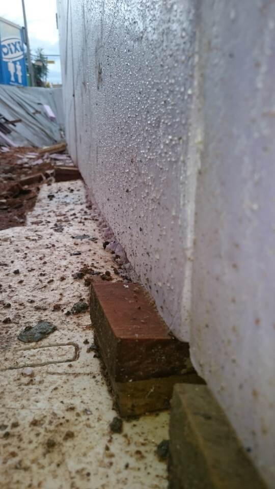 Außenbetonwand mit Feuchtigkeitsflecken; roter Backstein am Fundament, Schutt auf dem Boden.
