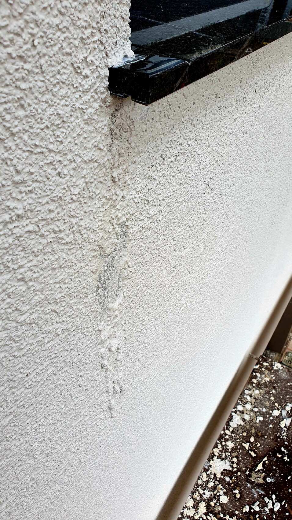 Außenwand mit grober Putzstruktur, schwarze Steinleiste oben, Feuchtigkeitsspuren und Staub am Boden.