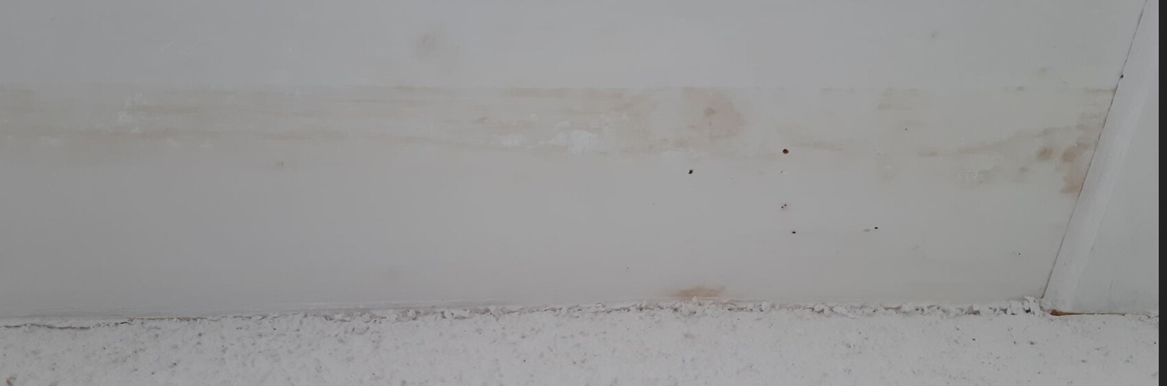 Weiße Wand mit einer horizontalen braunen Fleckenlinie und dunklen Flecken; unten rauer Boden.