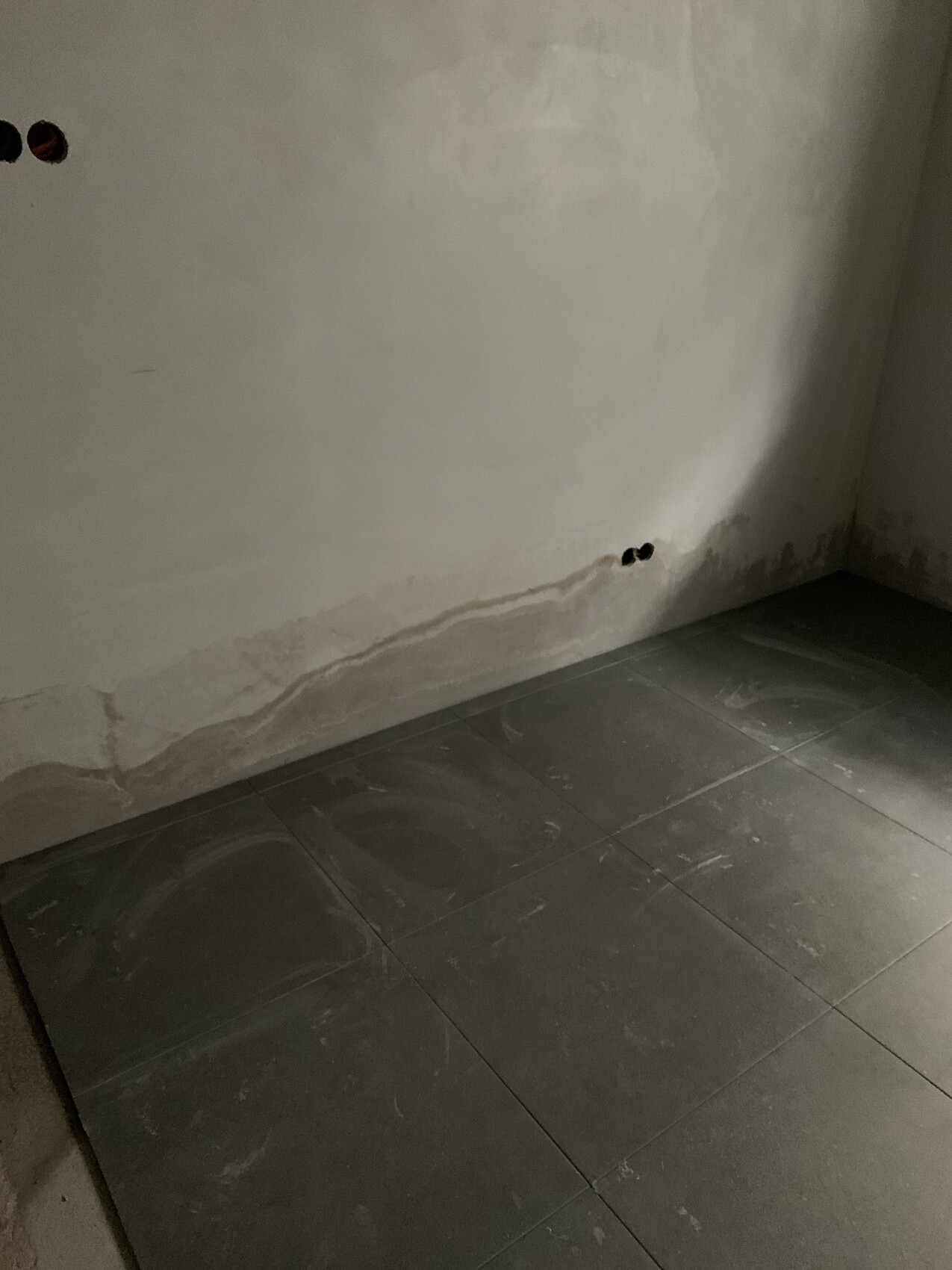 Ecke eines feuchten Raums mit rissiger Wand, Wasserflecken am Sockel und dunklen Fliesen.