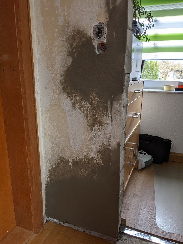 Wand mit grauem, unregelmäßigem Putz und freiliegter Elektro-Dose neben Holzschränken.