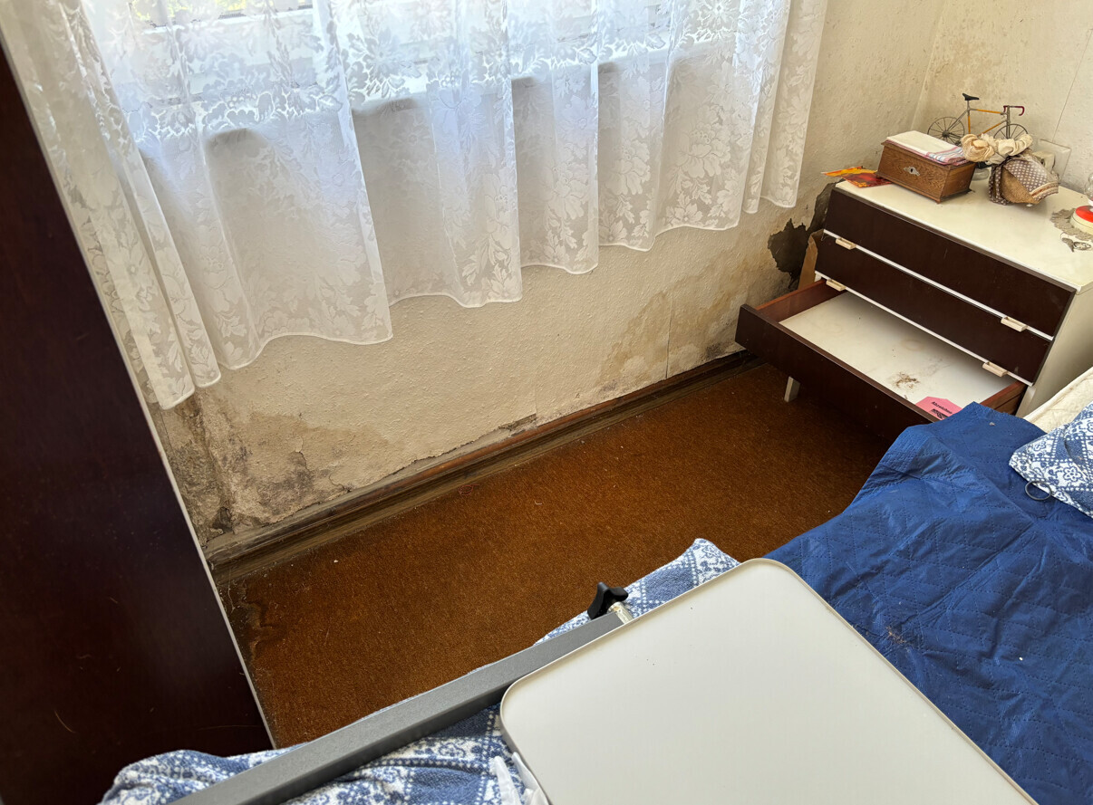 Schlafzimmer-Ecke mit nasser Wand, offener Kommode, Bett mit blauem Quilt und Spitzenvorhängen.
