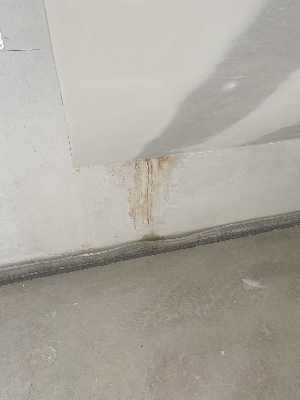 Braune Wasserflecken und Tropfen an einer Betonwand über dem Boden; Platte lehnt schräg an der Wand.