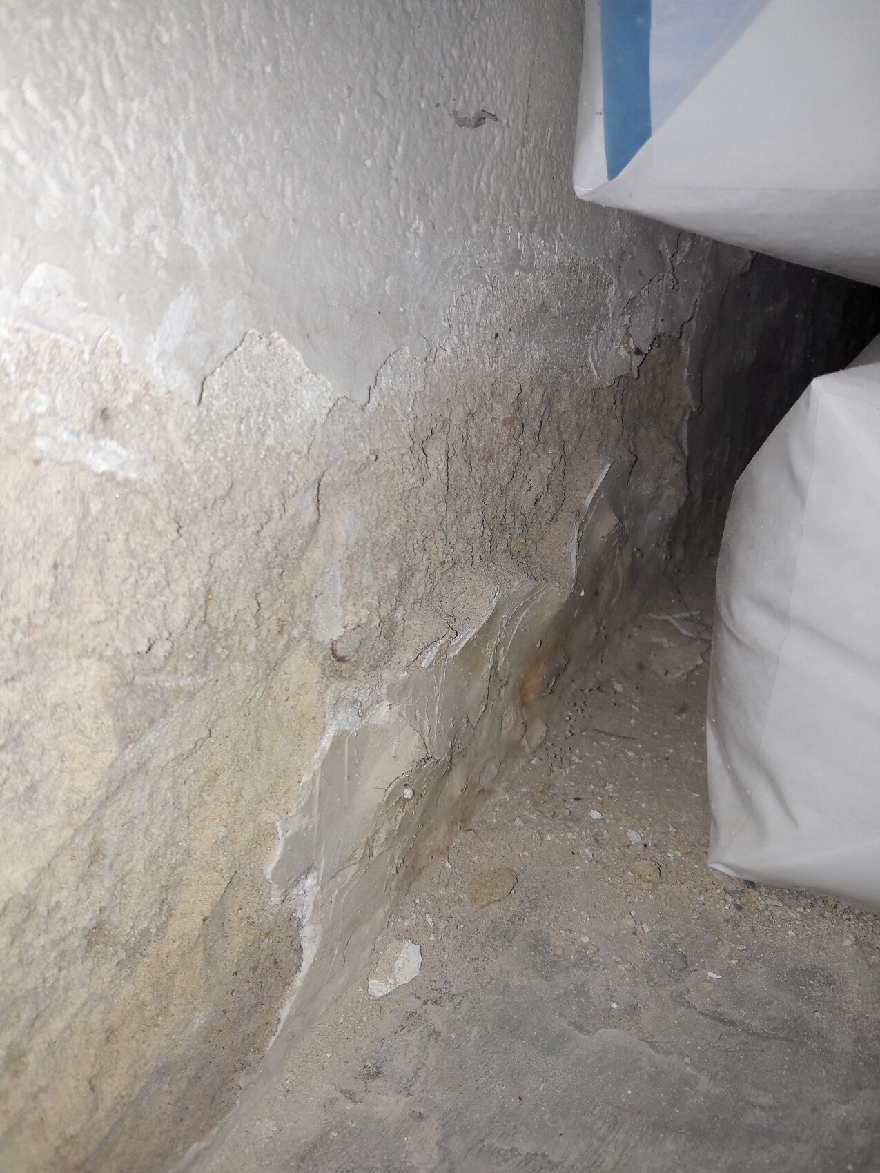 Kellerwand mit abgeplatztem Putz in der Ecke, staubig, Boden bröckelig, rechts liegt ein Sack.