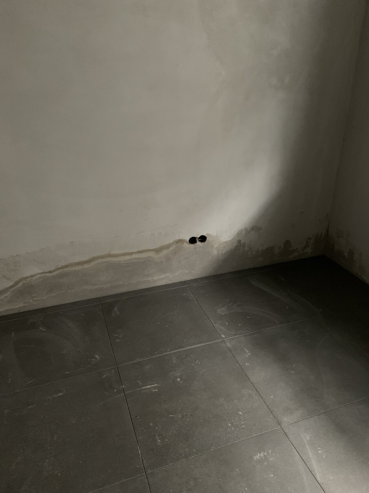 Wand-Ecke mit Feuchtigkeitsspuren, zwei runde Löcher, dunkle Bodenfliesen.