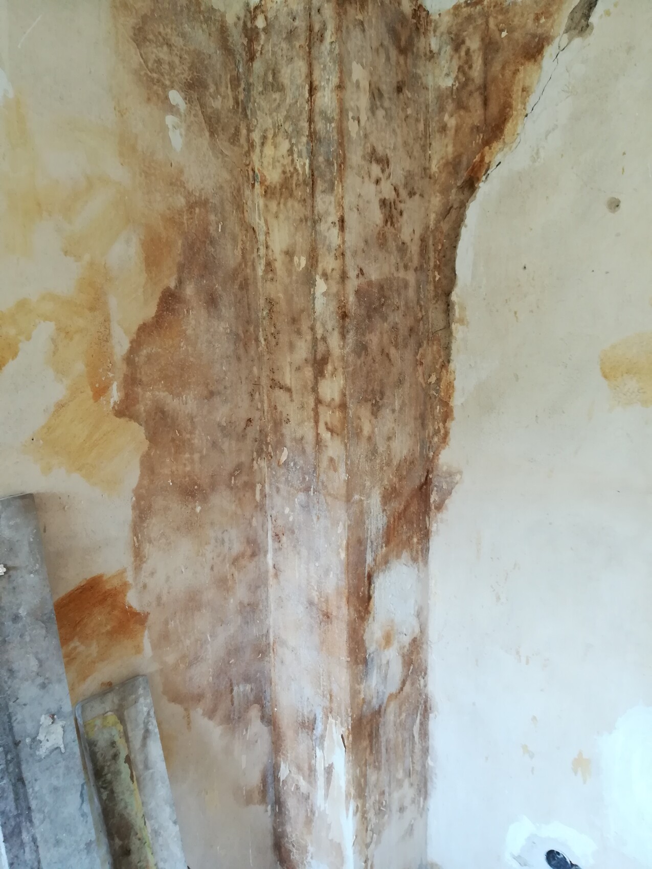 Feuchte Wand mit braunen Flecken und abgeblättertem Putz; Holzbretter lehnen links an der Wand.
