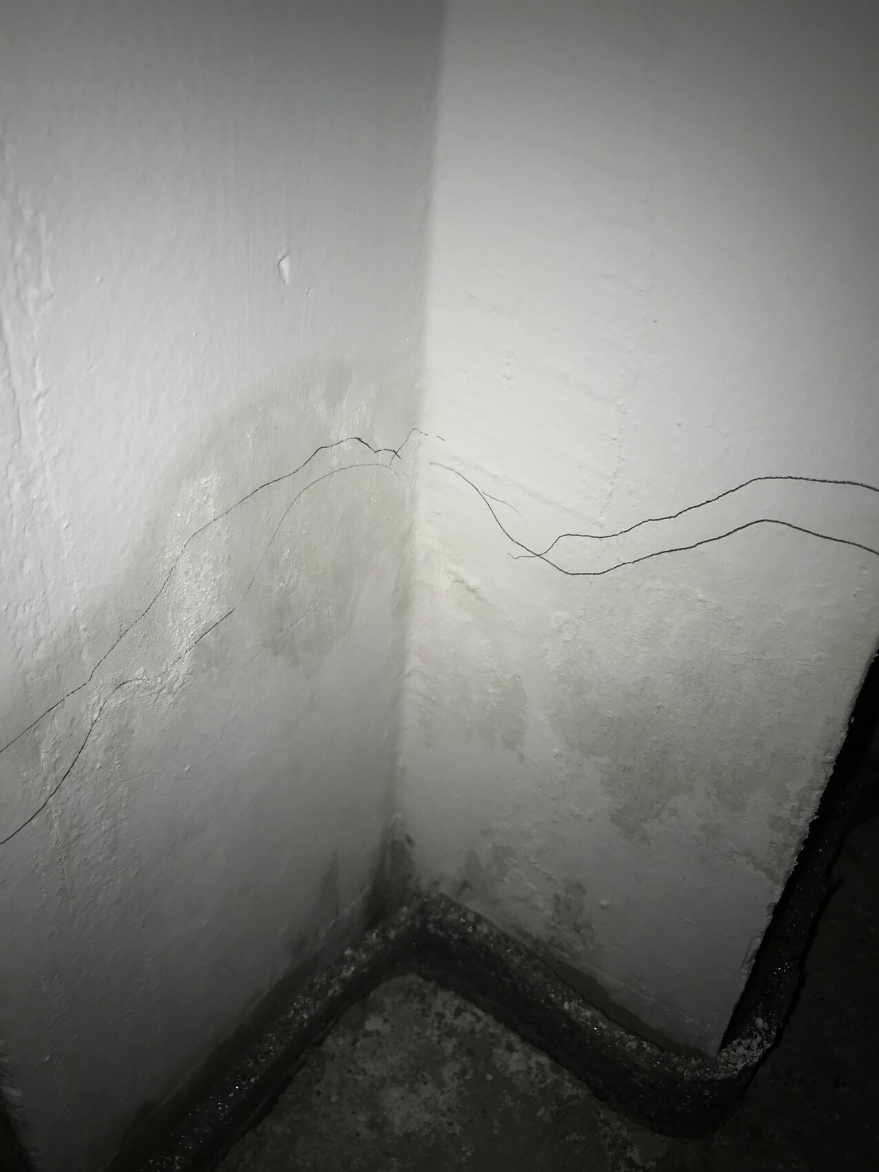 Eckansicht: weiße, rissige Wände mit feuchten Flecken am Boden; dunkles Rohr an der Ecke.