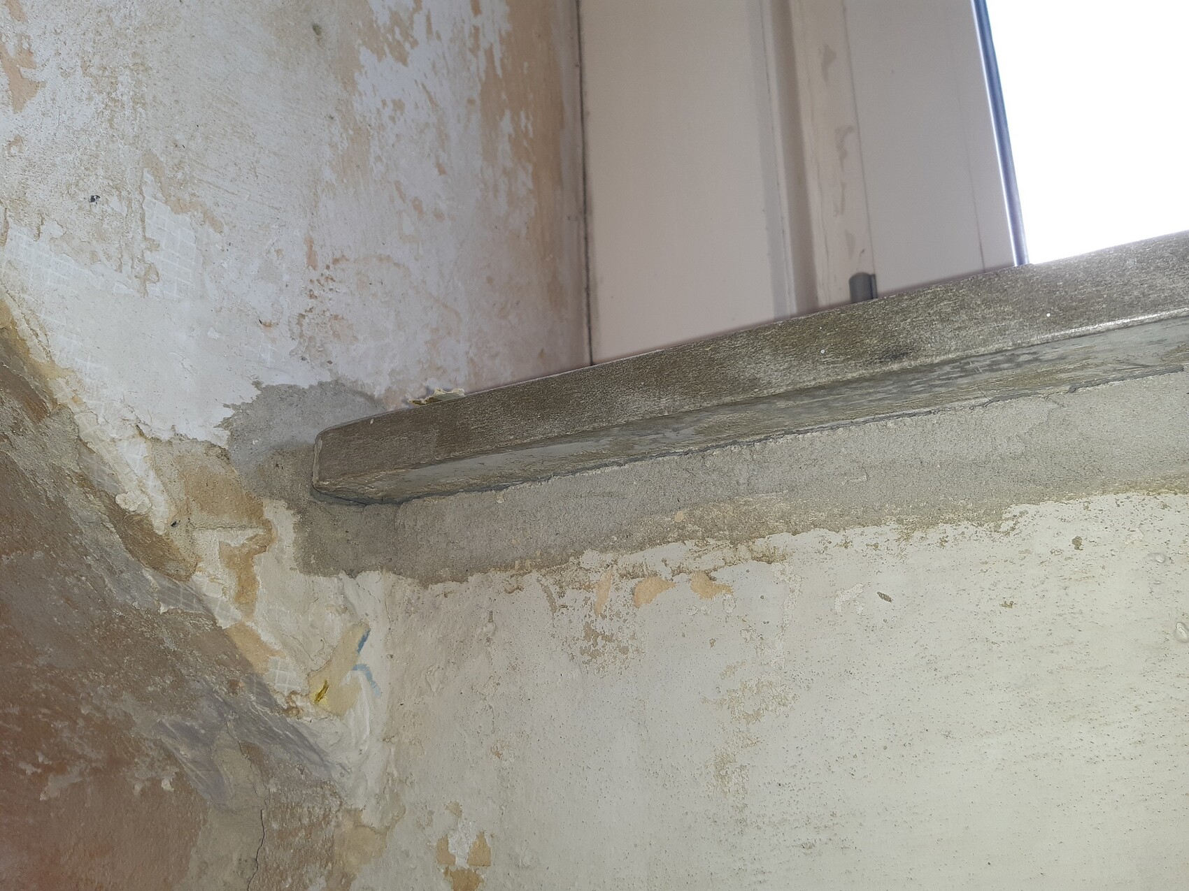 Rauer Wandabschnitt mit abblätterndem Putz; eine graue Betonleiste ragt als Fenstersims hervor.