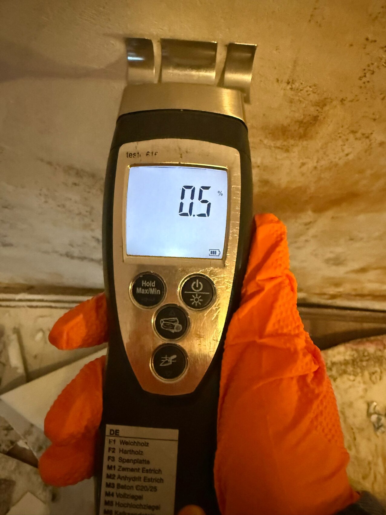 Hand mit orangefarbenem Gummihandschuh hält Feuchtigkeitsmessgerät an Betonboden; Anzeige 0,5%