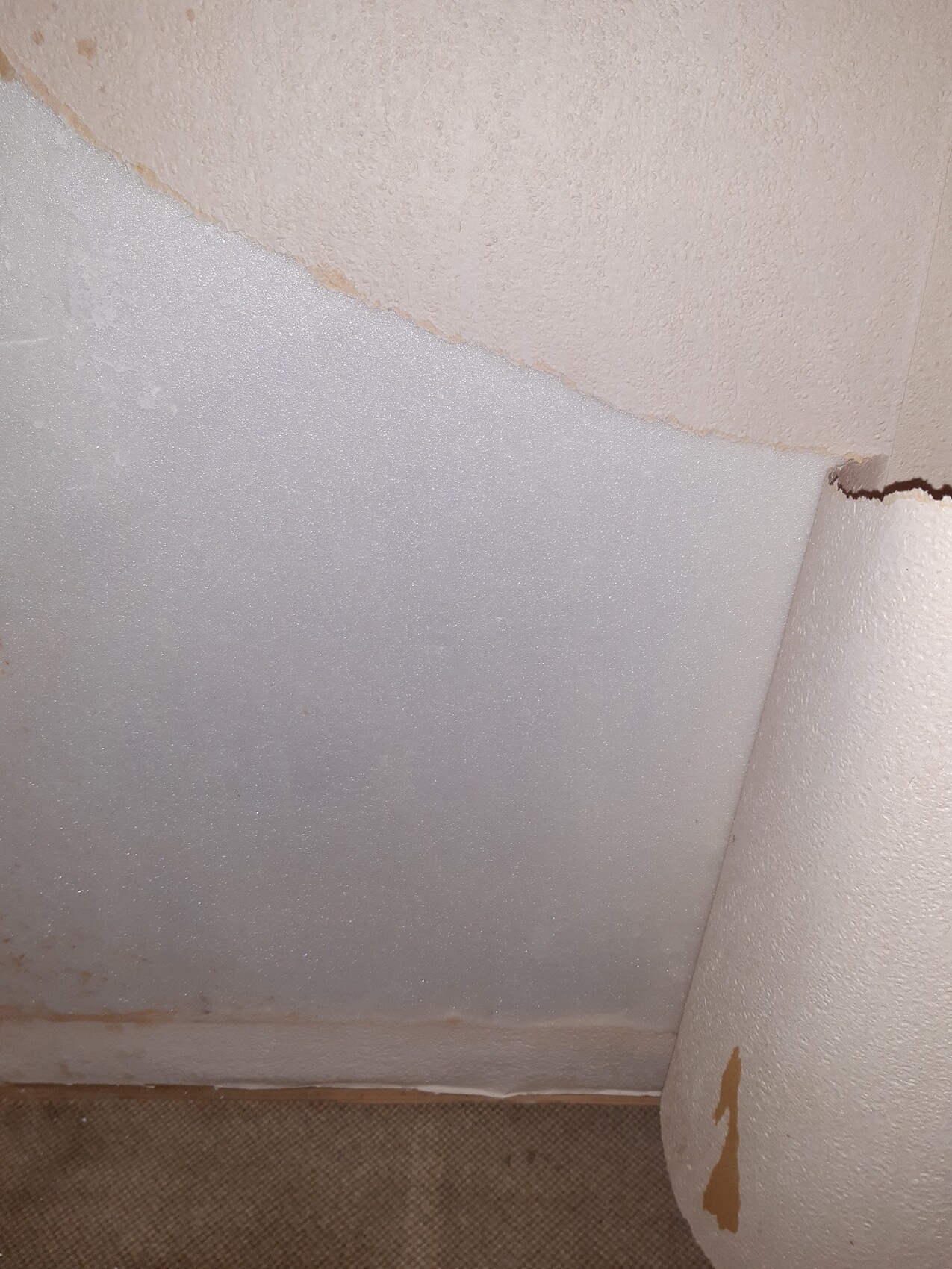 Innenraumecke mit rauem Putz, grauer Schaumstoffplatte und abblätternder Farbe an Säule, Teppichboden.