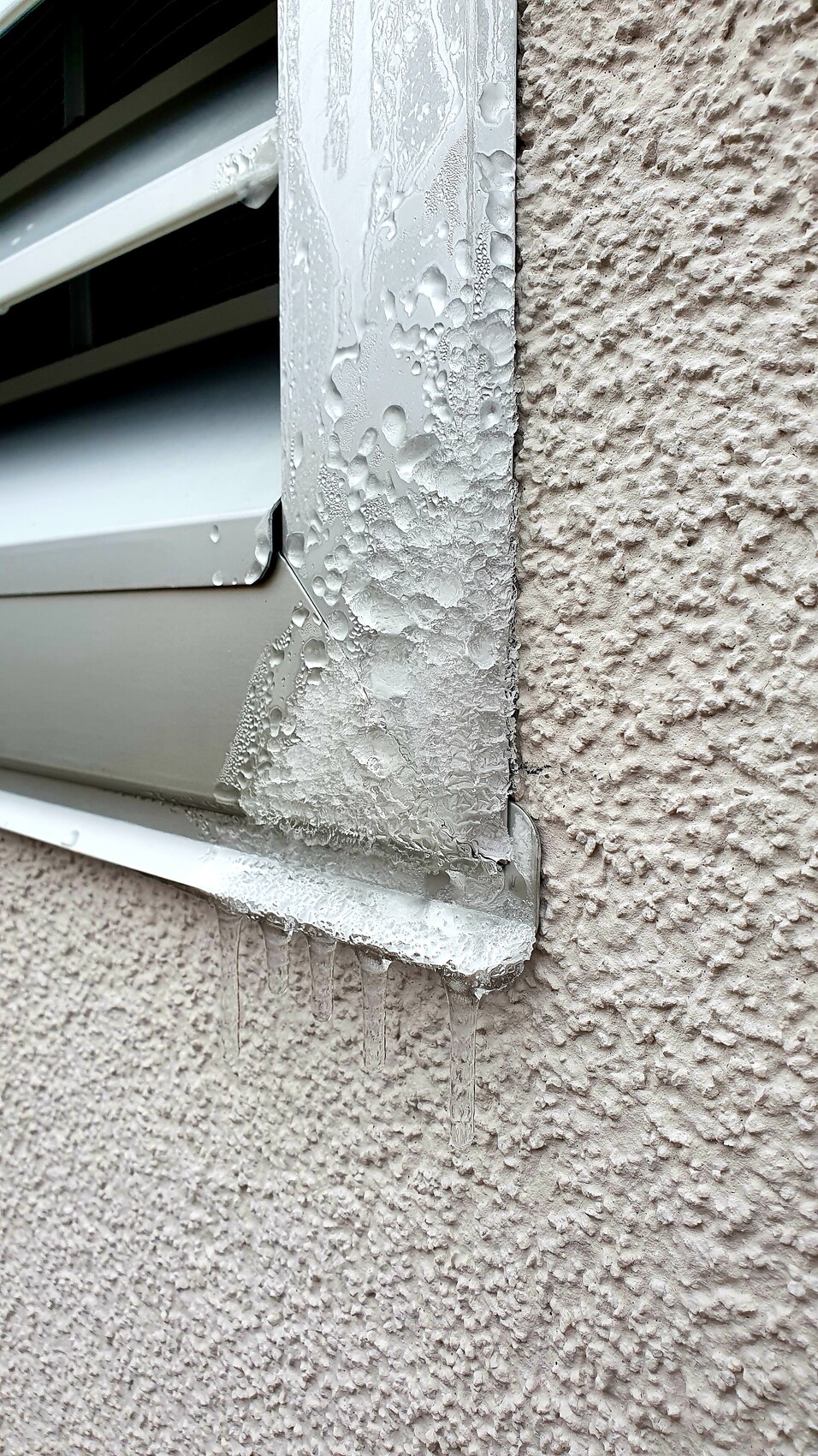 Außenfensterrahmen aus Metall mit Wassertröpfchen und Eiszapfen am rauen Putz der Wand.