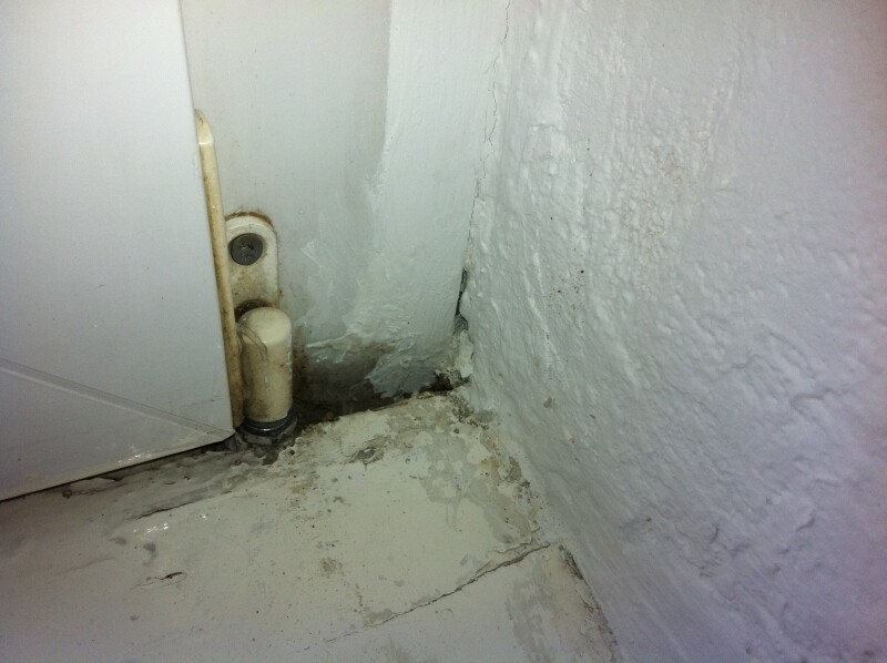 Ecke eines Raums mit weißer Wand, feuchtem Boden und rohrförmigem Abfluss.