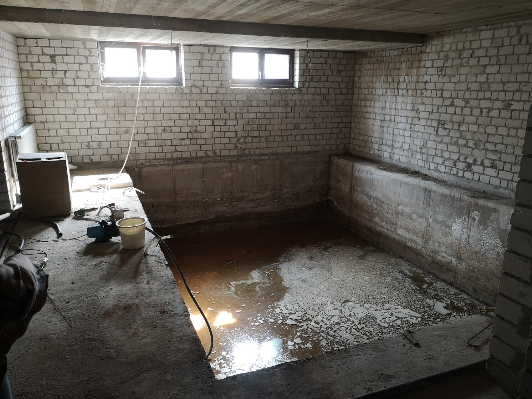 Kellerraum mit einem ausgehobenen Becken, braunem Wasser, Ziegelwänden und kleinem Fenster.