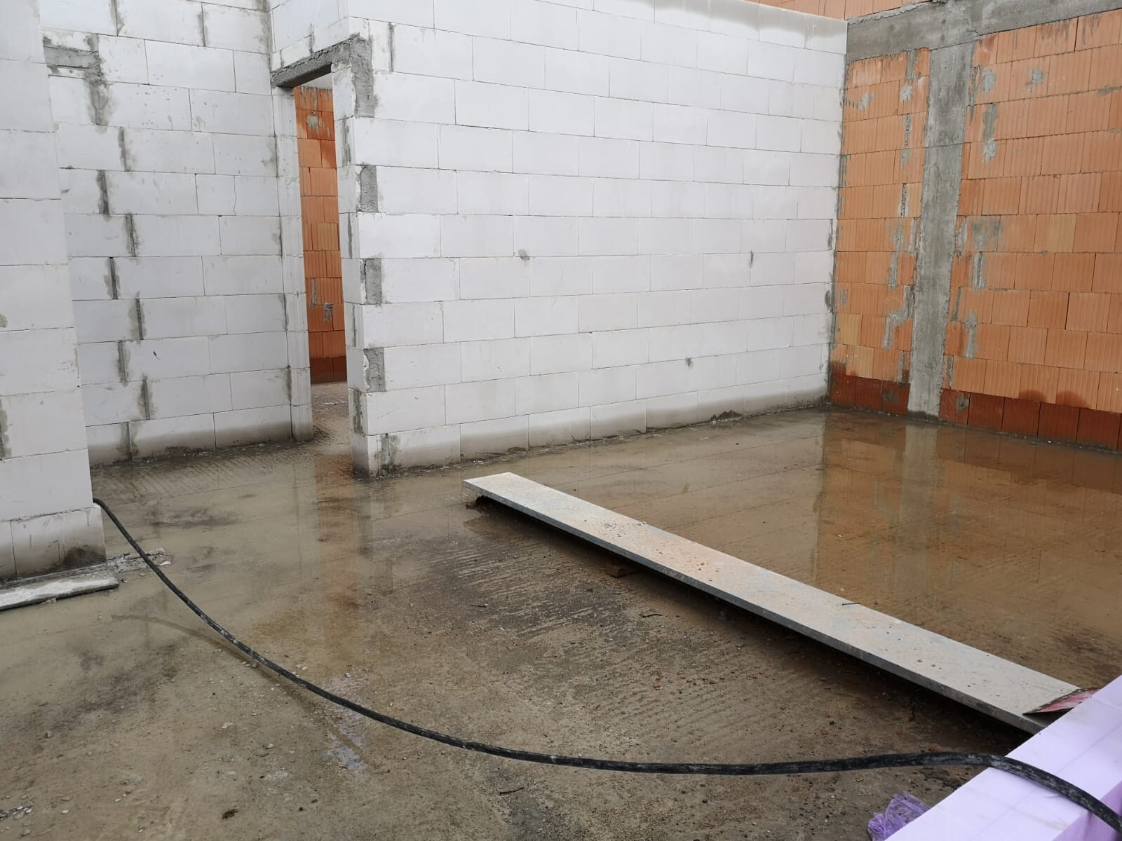 Baustelle: Innenraum mit weißen Blockwänden, rechte Ziegelwand, nasser Boden und Planke.