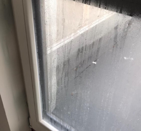 Fensterrahmen links, beschlagene Glasscheibe mit Regentropfen; graue Außenansicht.