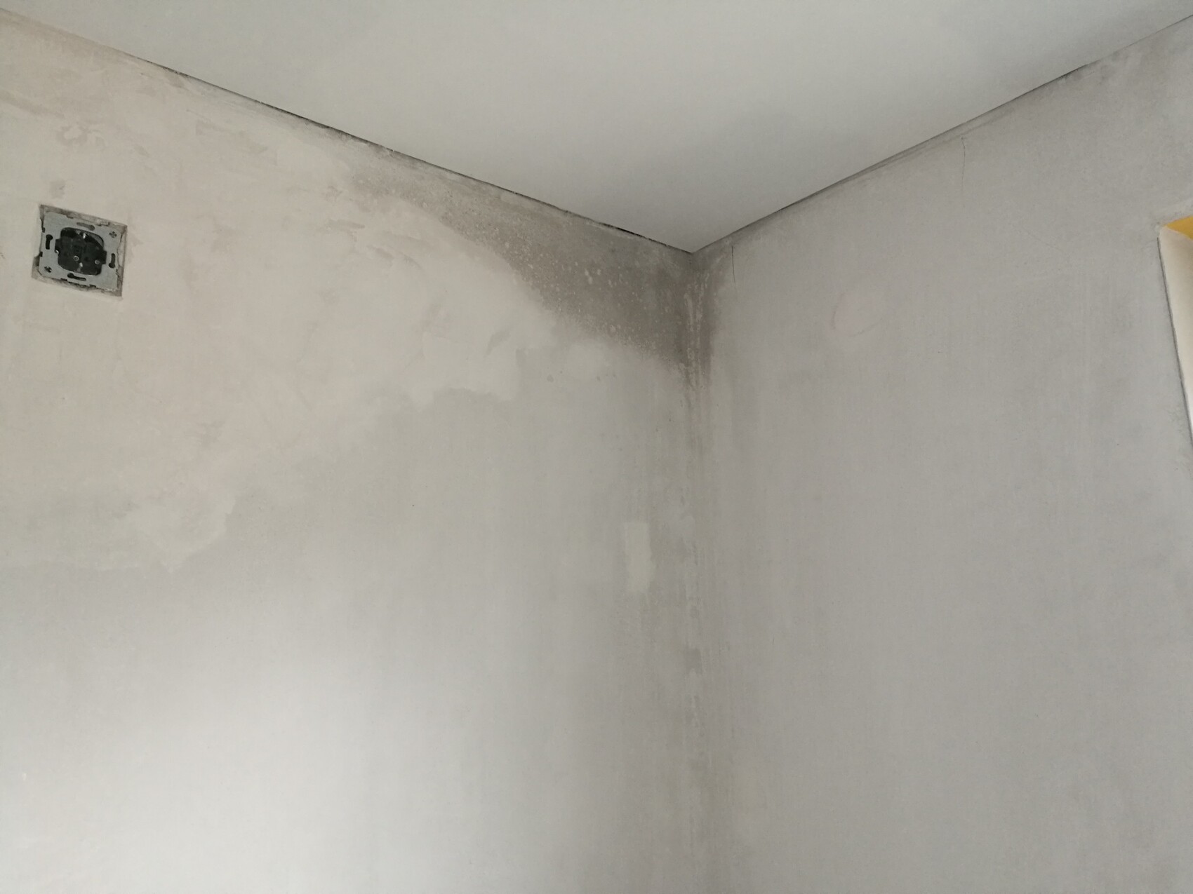 Feuchtigkeitsschaden in Innenraum-Ecke, nasse Flecken an Wand und Decke, Steckdose sichtbar.