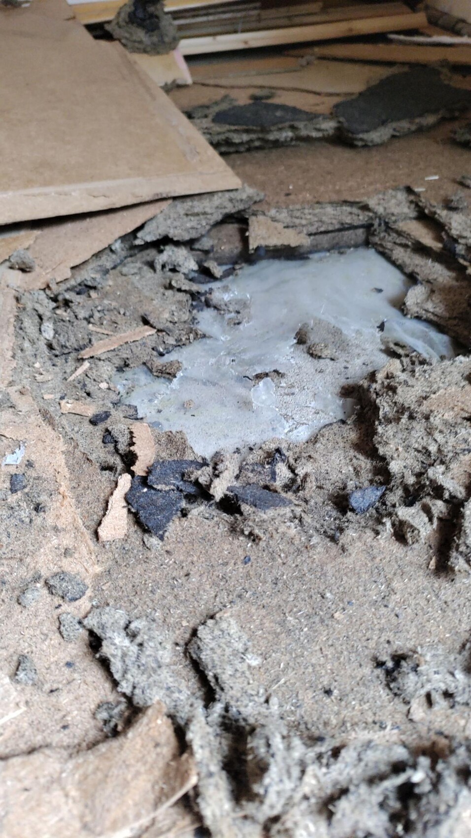 Beschädigte Bodenfläche mit bröckelndem Schutt, Holzplatten und einer grauen Pfütze.