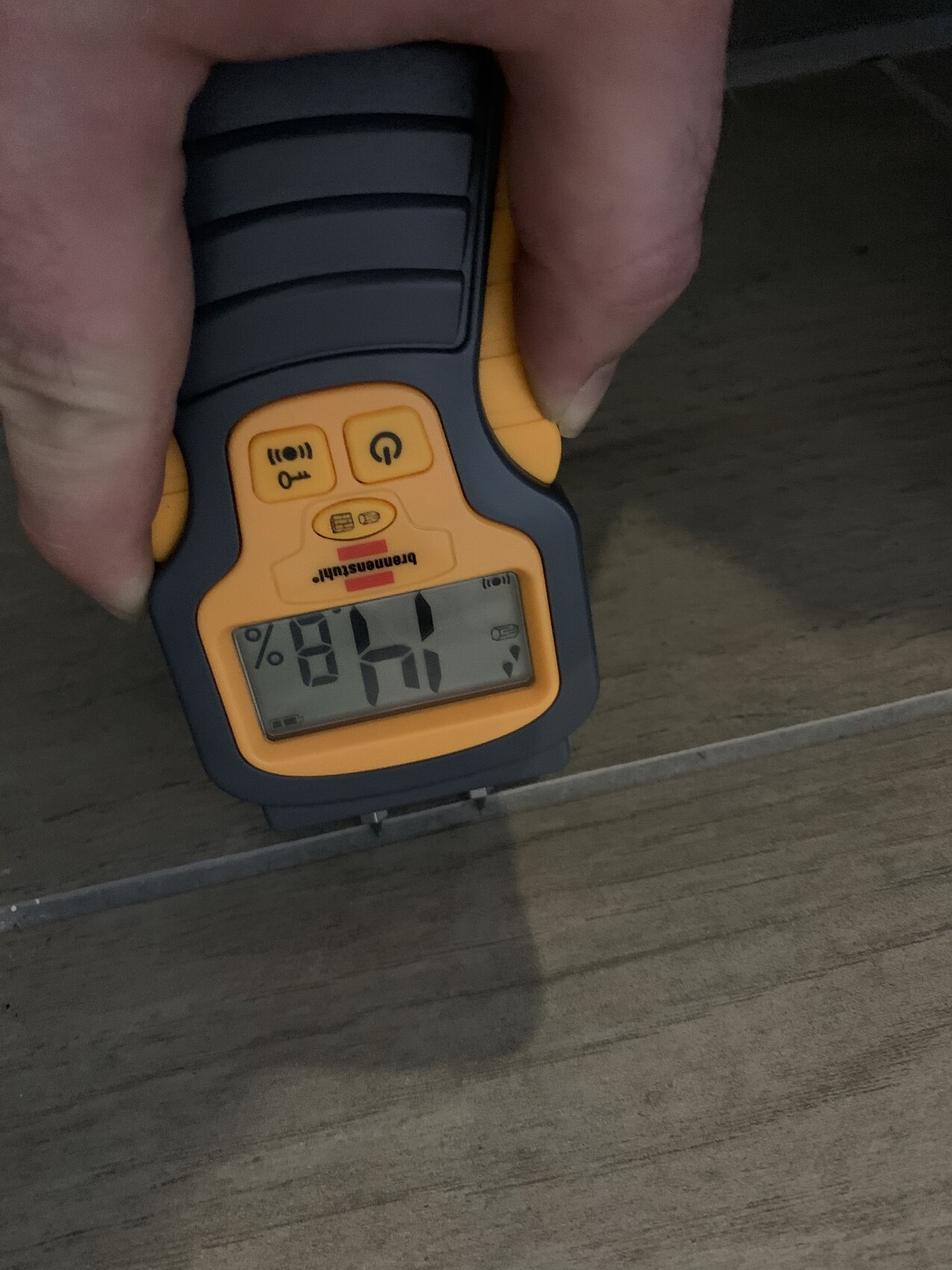 Hand hält orangefarbenes Feuchtigkeitsmessgerät an Holzboden; Display zeigt Feuchtigkeitswert in Prozent.