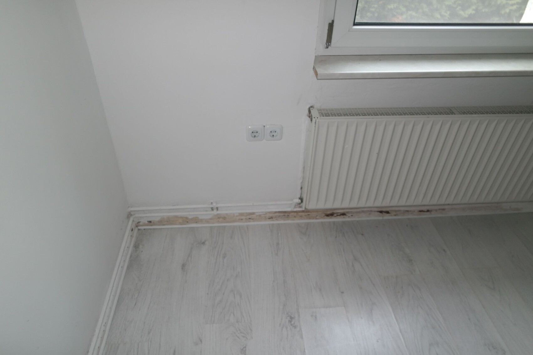 Weißes Zimmer-Eck mit Fenster über Heizkörper; zwei Steckdosen; beschädigte Bodenleiste.