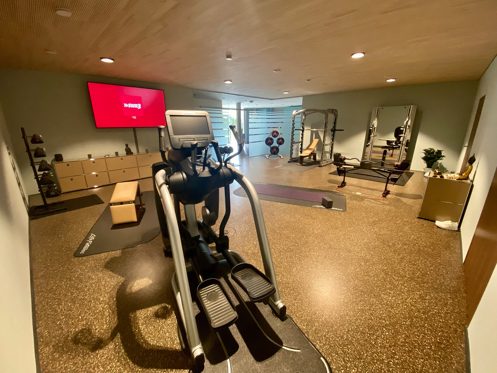 Crosstrainer im Fitnessraum, Hantelbank, Spiegelwand, Gewichte, TV an der Wand.