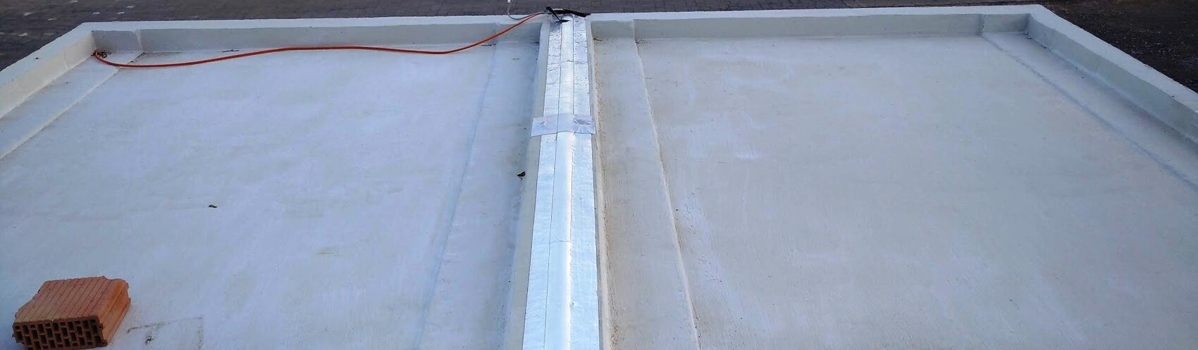 Weißes Flachdach mit zentralem Metallstreifen, rotes Kabel links, Backstein unten links.