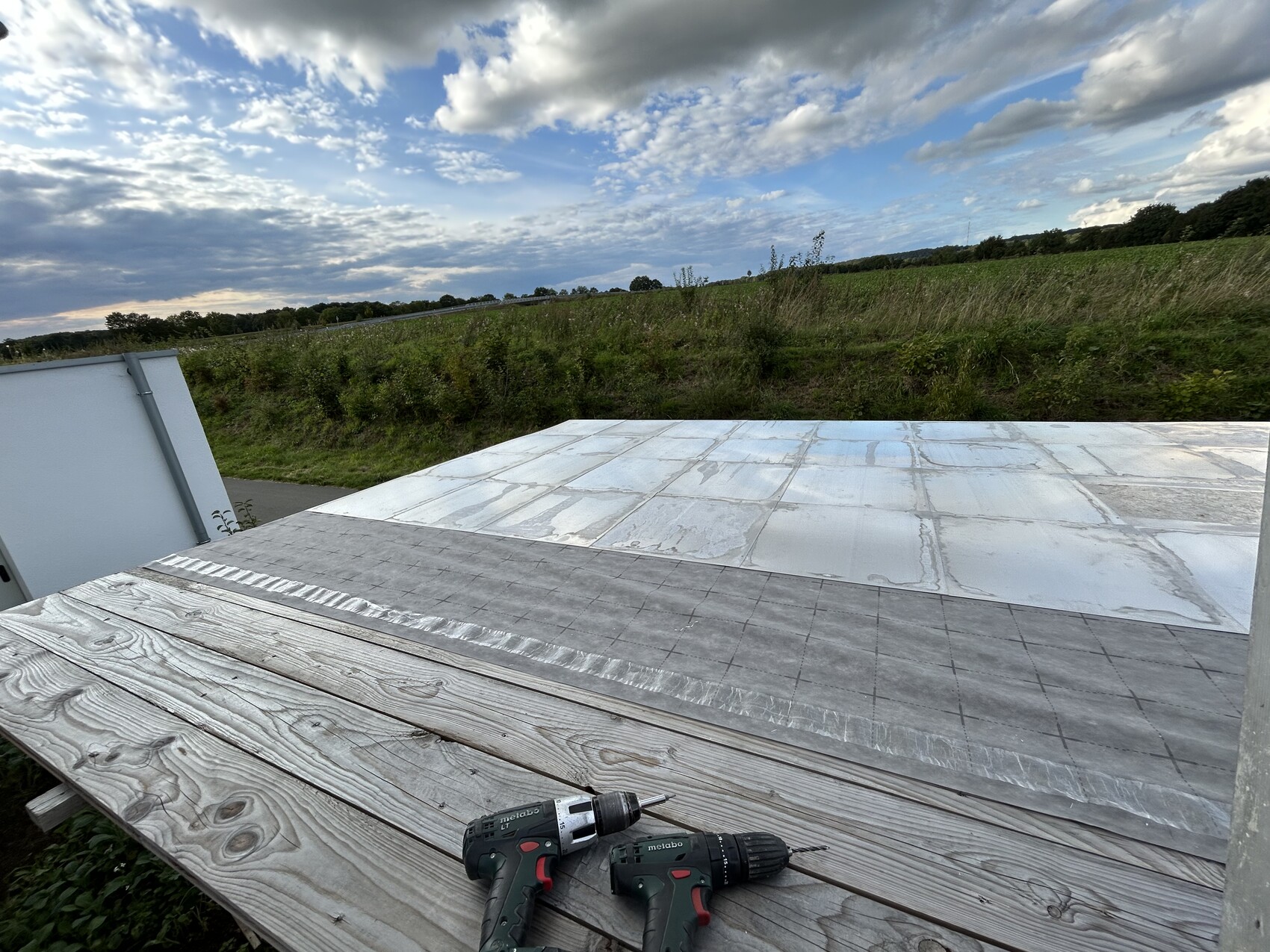 Holzdeck mit zwei Bohrmaschinen vor flachem Metalldach; Felder und Wolkenhimmel im Hintergrund.