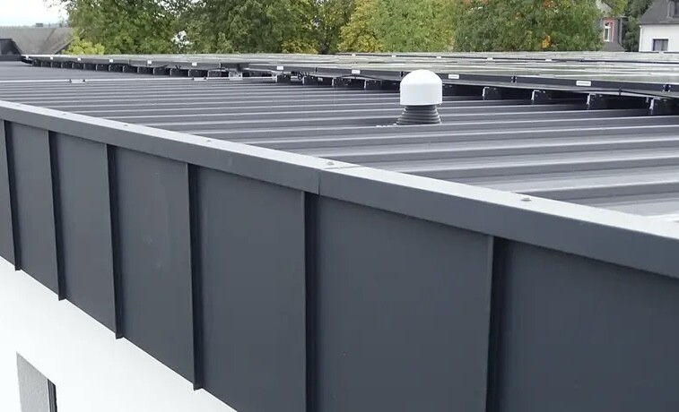 Graues Flachdach aus Metallpaneelen mit weißem Dachlüfter; Hintergrund mit Bäumen.