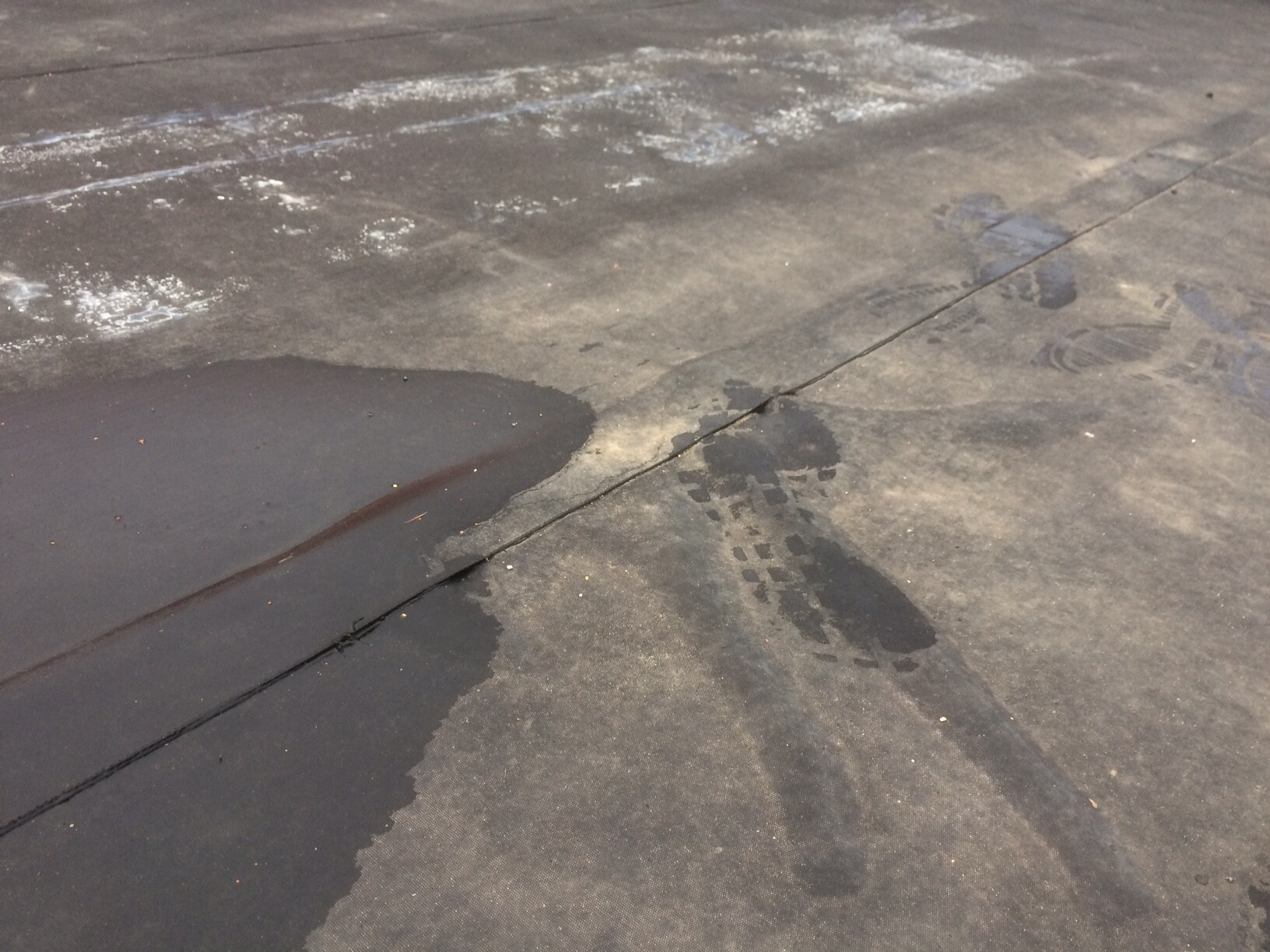 Nasse Asphaltfläche mit Pfütze links, dunkle Flecken und Schuhabdrücke rechts.