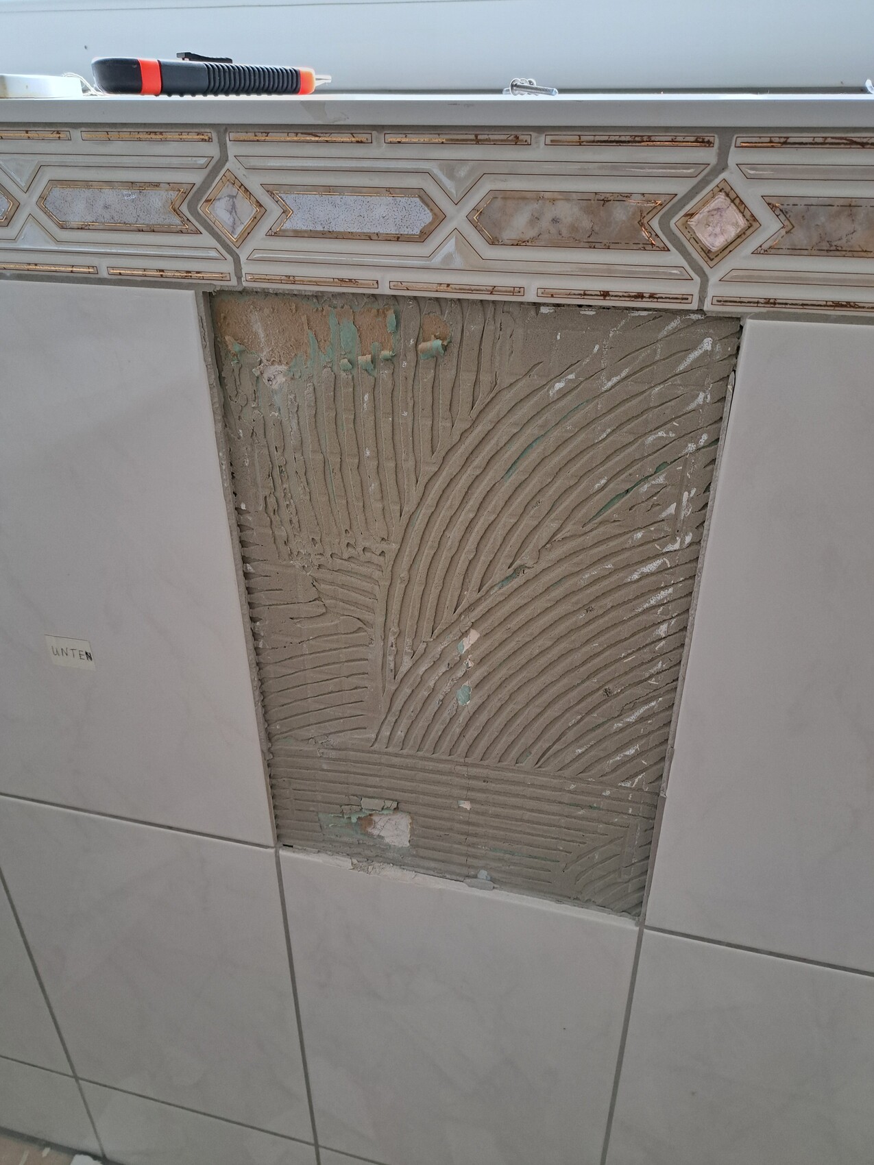 Fliesenwand mit freigelegtem Mörtelbereich, Werkzeug oben, dekorative Bordüre.