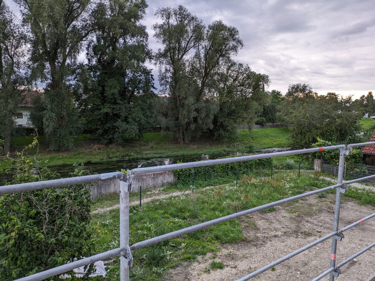 Kanal mit Ufergras und großen Bäumen, Geländer im Vordergrund, ruhige ländliche Szene.