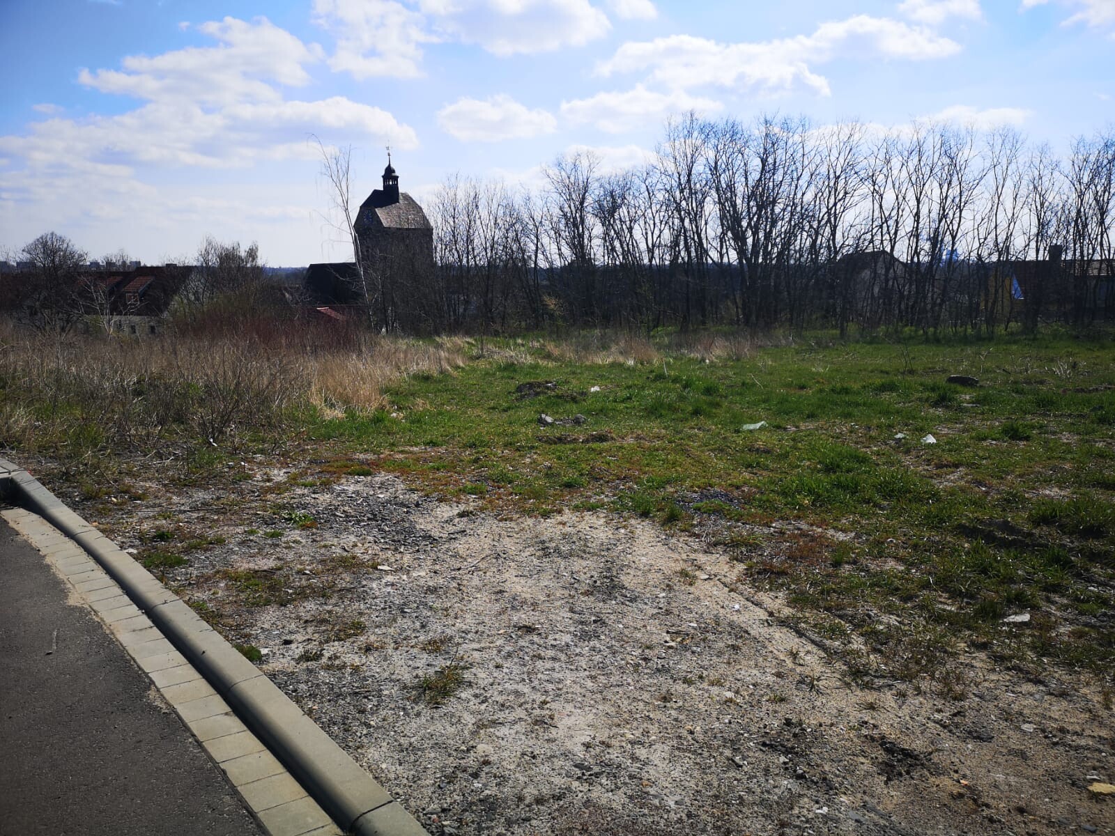 Leeres Grundstück mit Kiesboden, Grasflächen, Baumreihe und Kirchturm im Hintergrund