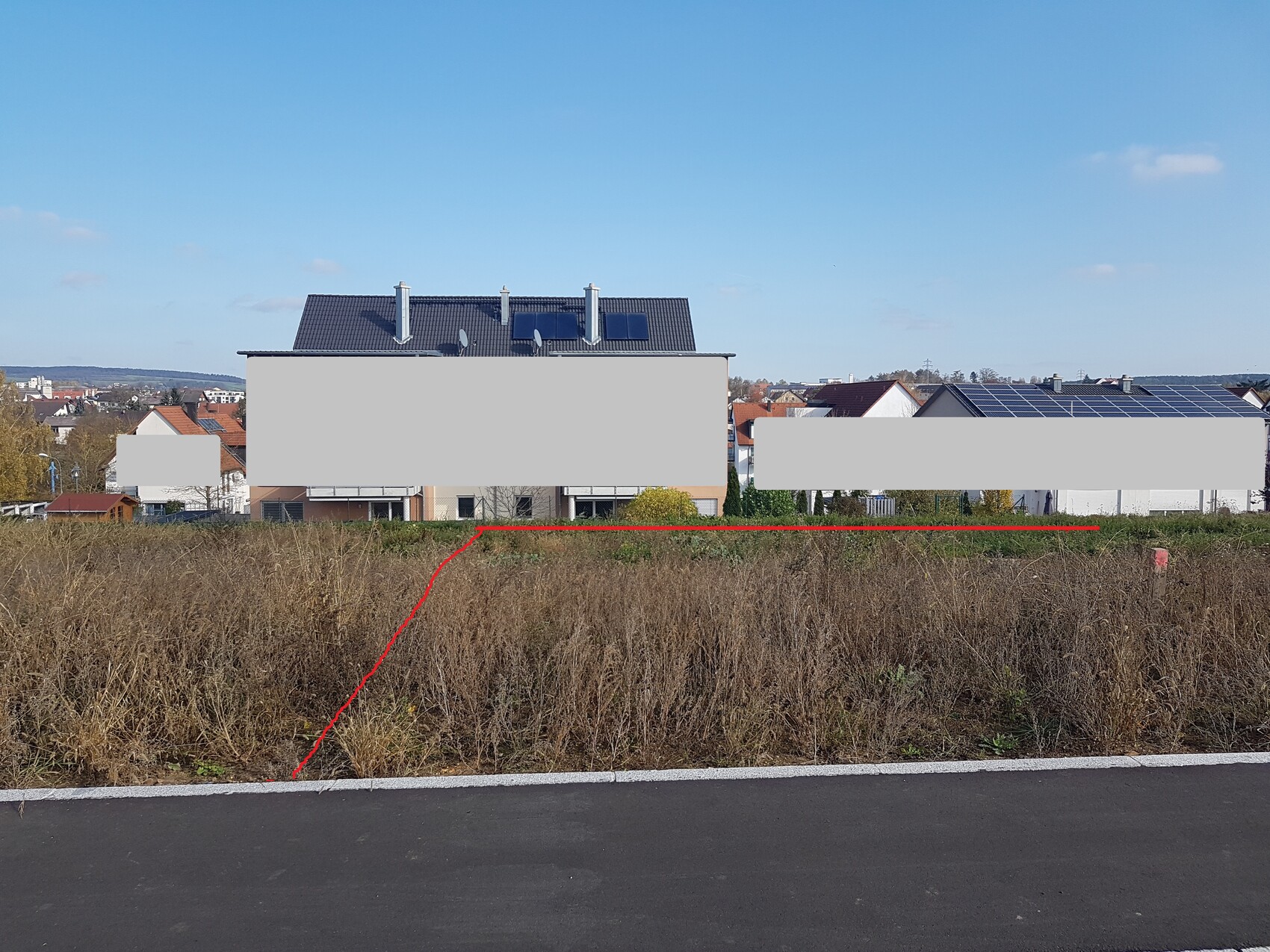 Grasbewachsener Bauplatz vor Wohnhäusern mit Solarzellen auf Dächern; roter Weg markiert den Zugang im Vordergrund.