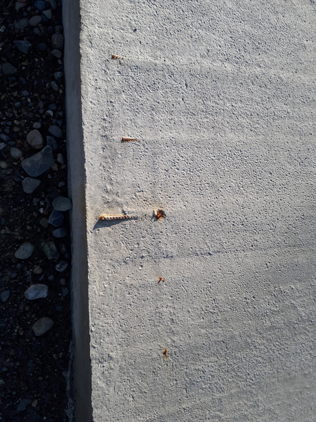 Hellgraue Betonplatte mit rostigen Nägeln, daneben Kieselsteine im Schatten.