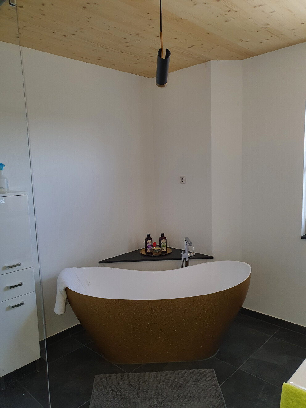 Freistehende braune Badewanne mit weißem Innenraum in modernem Bad, Holzdecke und Pendelleuchte.