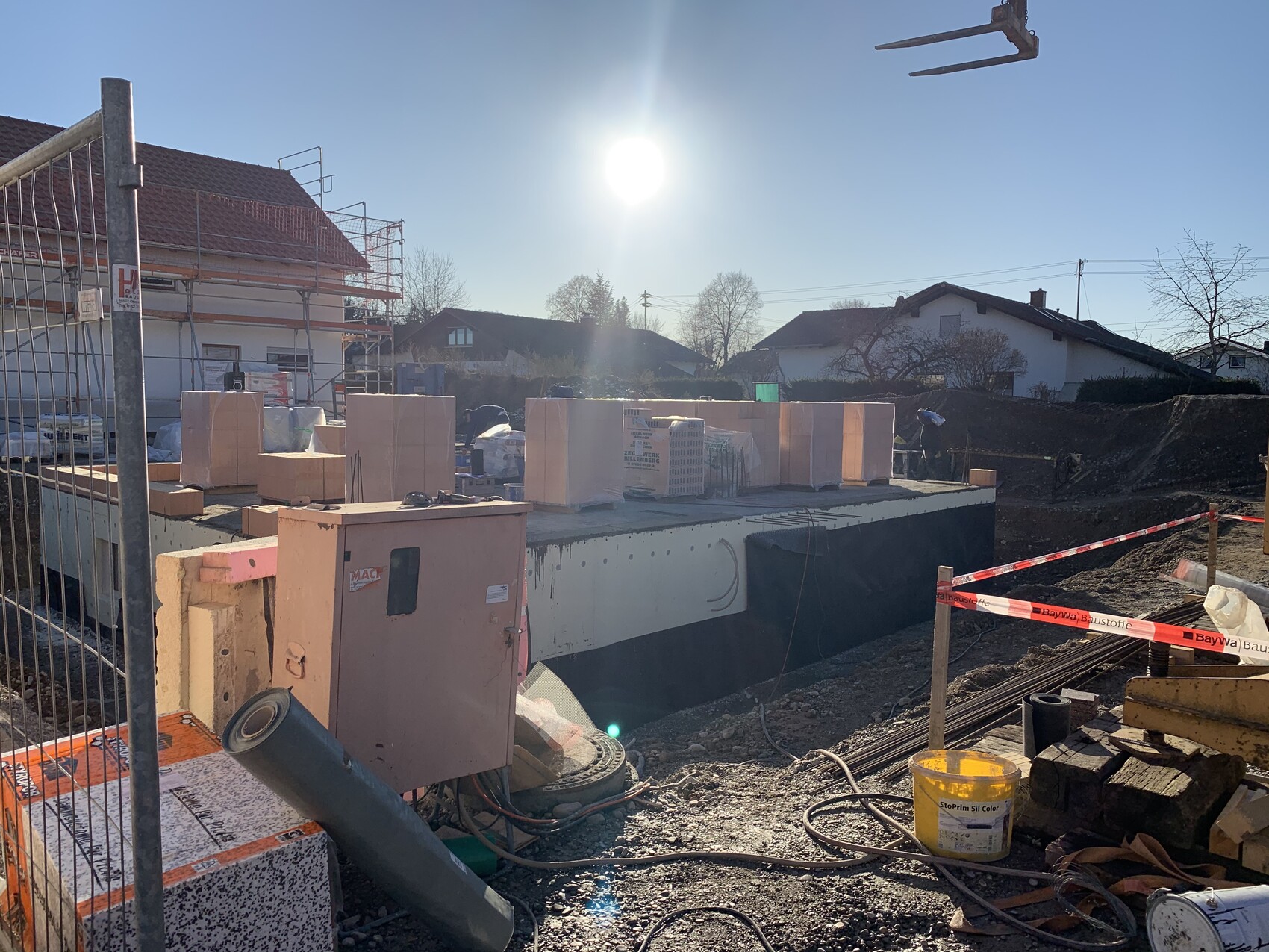 Baustelle mit Fundamentplatte, gestapelten Blöcken, Gerüst am Haus, Sonne am Himmel.