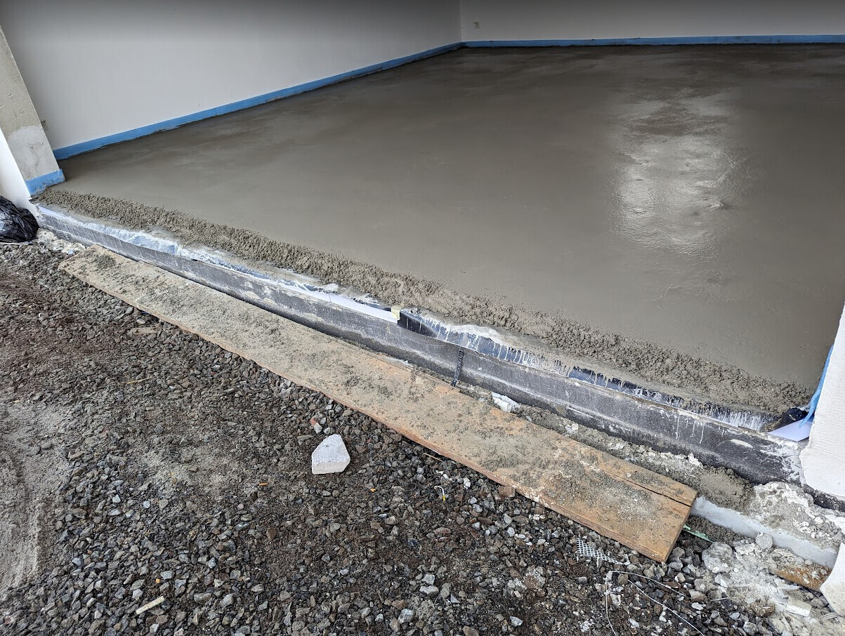 Frisch geglättete Betonbodenplatte in Bauphase; Innenraum, daneben Kies und Holzrampe.