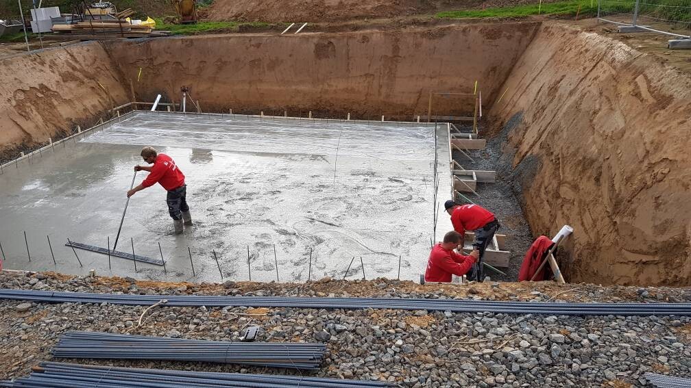 Drei Bauarbeiter in roter Kleidung arbeiten in einer Baugrube an einem Betonfundament mit Bewehrung.
