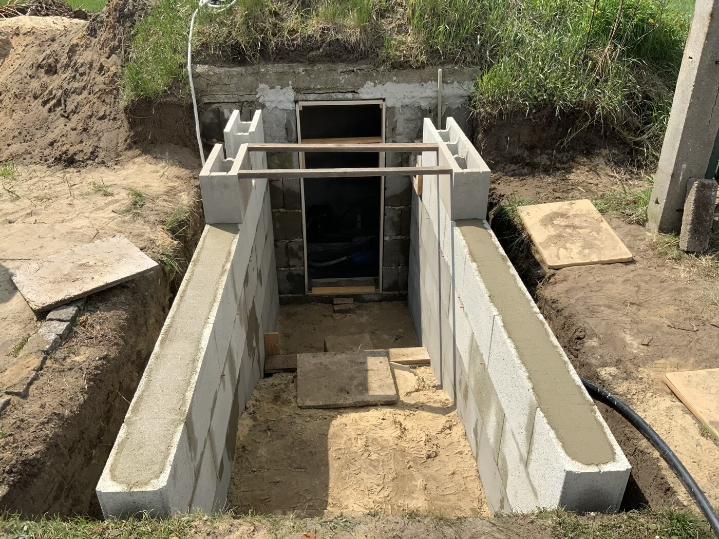 Kellerabgang aus Betonblöcken mit Holzbalken-Brücke über Baugrube.