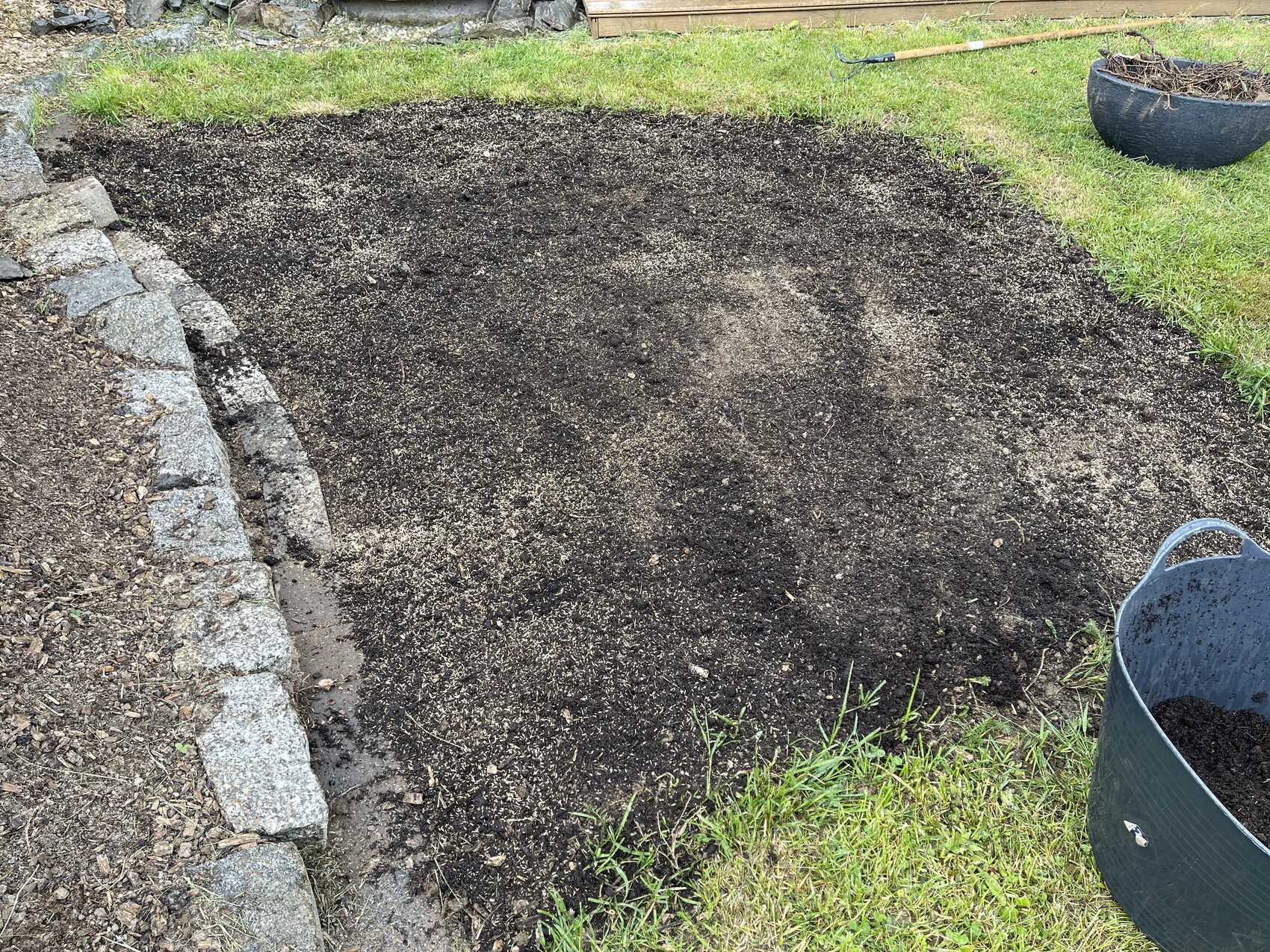Ovales Beet aus dunkler Erde im Garten, links Steinmauer, Grasrand, Rechen oben.