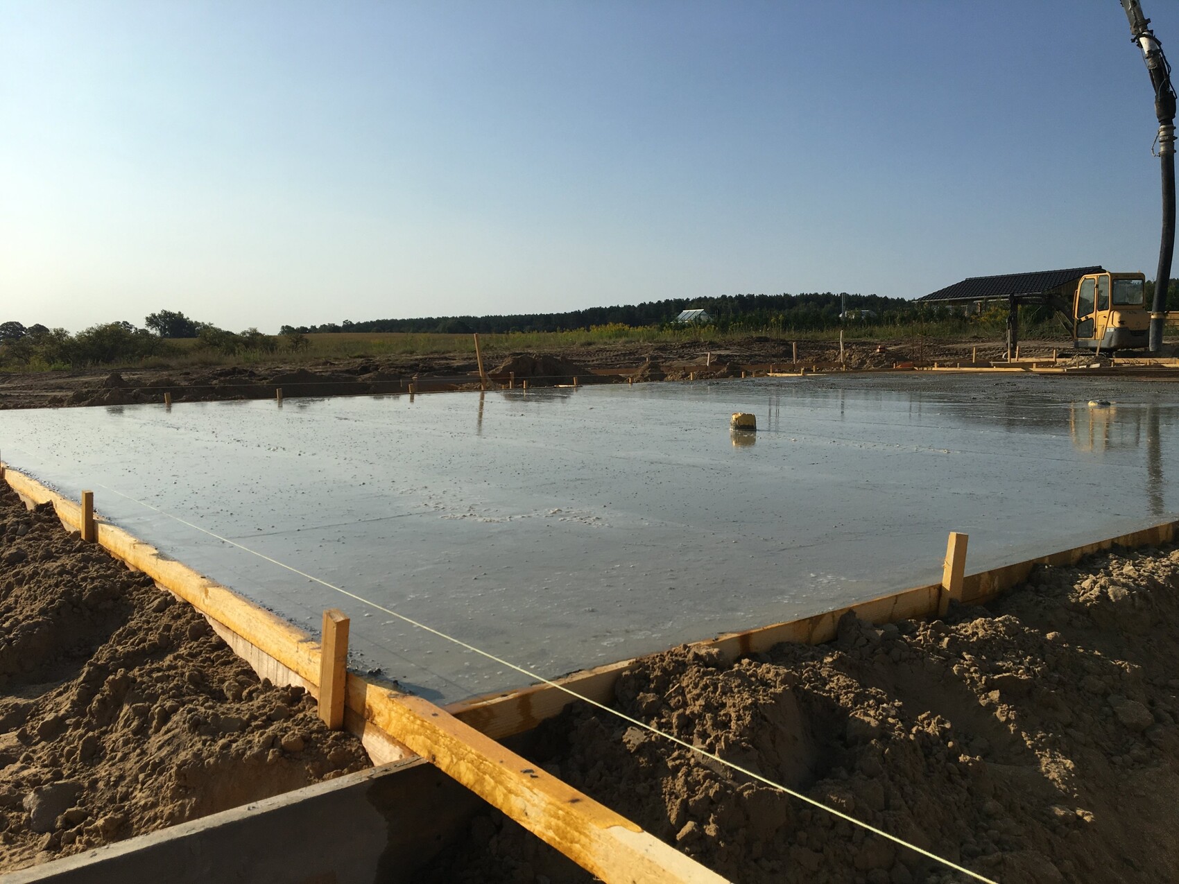 Frisch gegossene Betonplatte in Holzfundamentrahmen auf Baustelle, Baufahrzeug rechts.