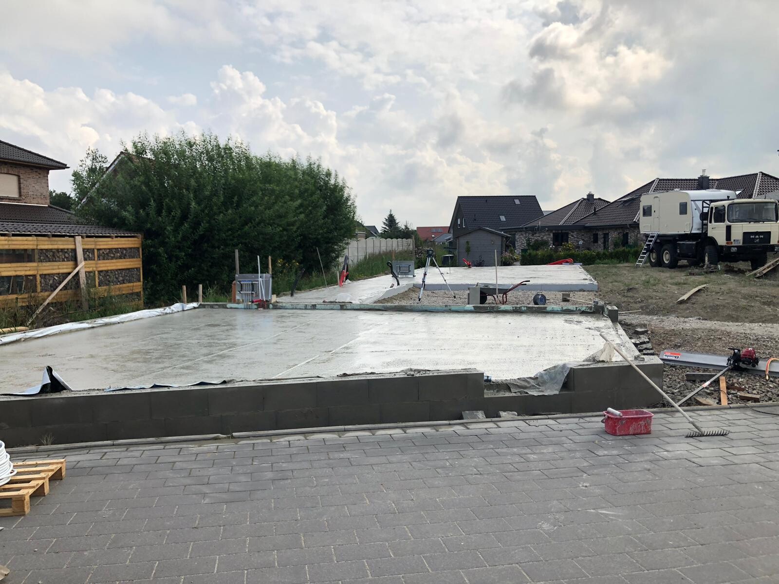 Frisch gegossene Betonplatte auf Baustelle mit Werkzeugen, Lastwagen und Häuser im Hintergrund.