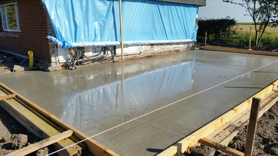 Frisch gegossene Betonplatte auf Holzformrahmen; Haus mit blauer Plane im Hintergrund.