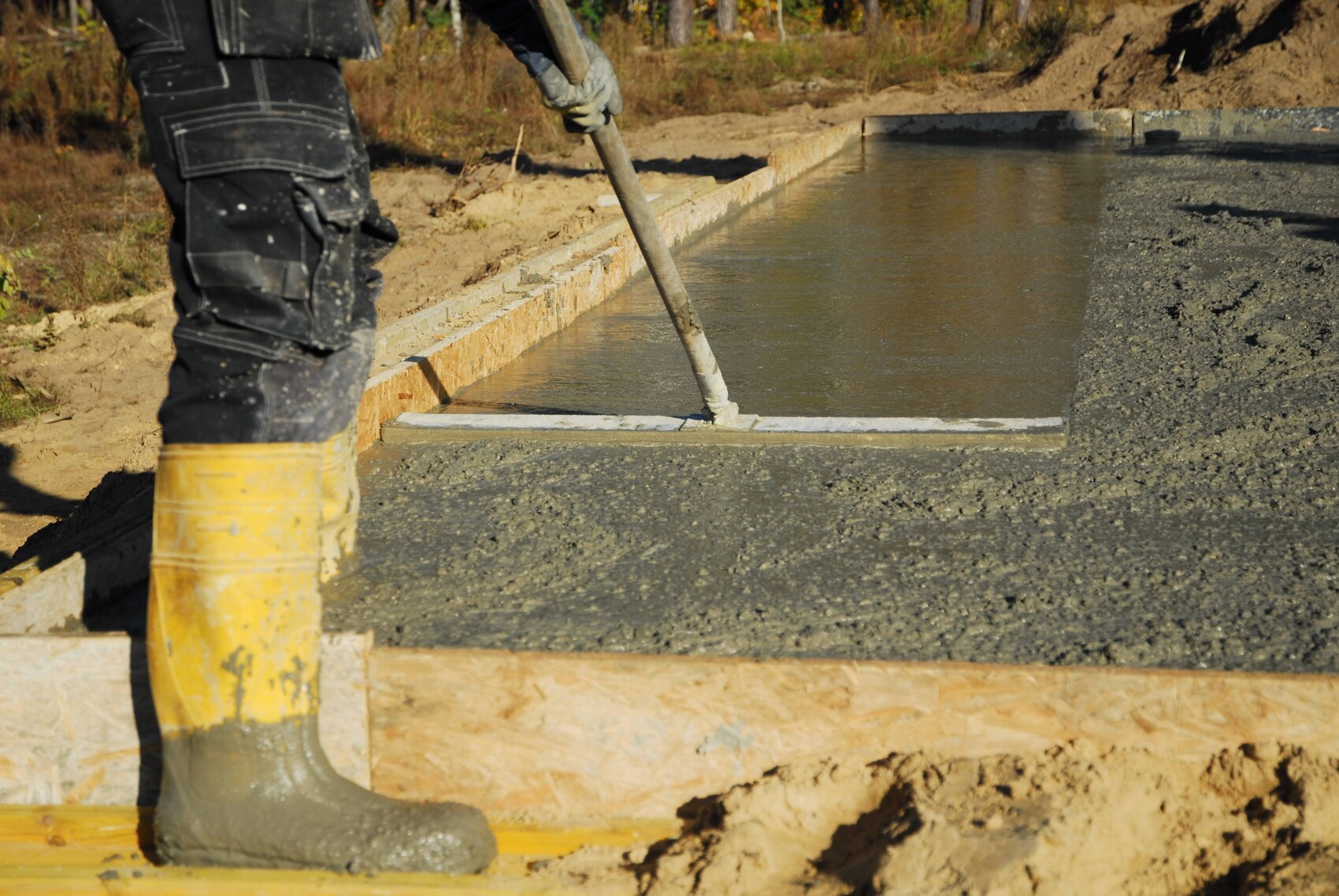 Arbeitskraft glättet frischen Beton in einer Holzform; gelbe Gummistiefel im Vordergrund.