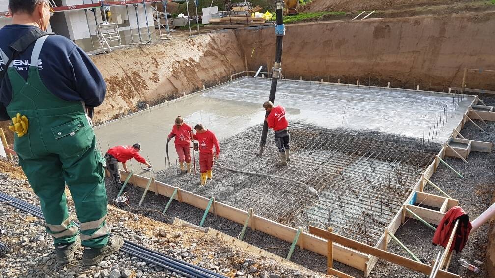 Vier Bauarbeiter gießen Beton in eine Baugrube, Bewehrung liegt bereit, Pumpenschlauch sichtbar.