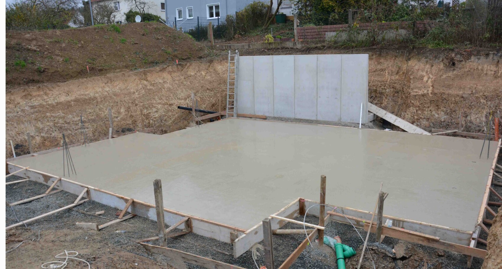 Frisch gegossene Betonsohle in einer Baugrube mit Holzformen und Leiter, Häuser im Hintergrund.