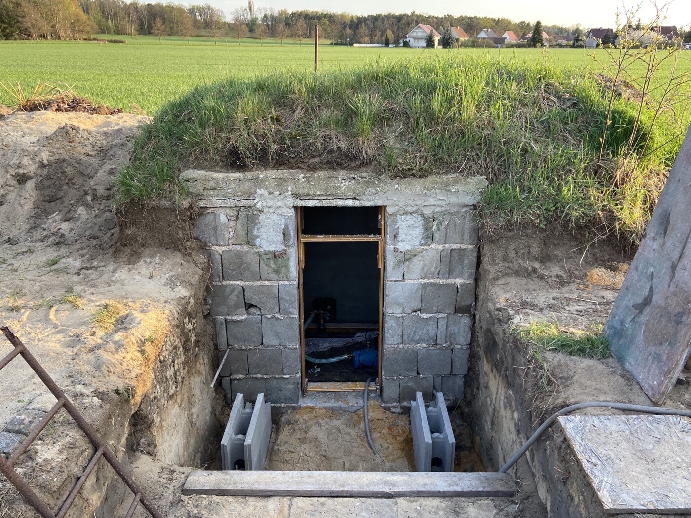 Unterirdischer Eingang aus Betonblöcken im Erdhügel, offen, Feld und Häuser im Hintergrund.