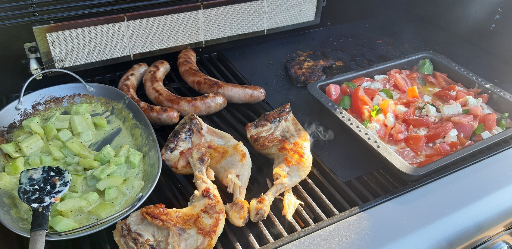 Grillplatte mit Würsten, Hähnchenkeulen; links Gurken in Dip, rechts Tomaten-Paprika-Gemüse.