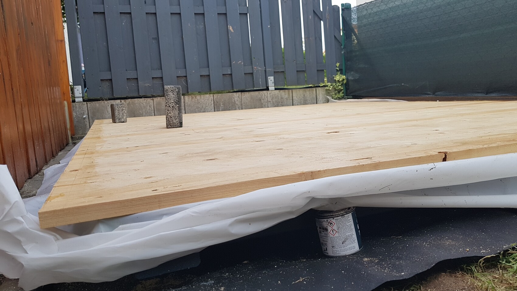 Provisorische Holzplattform im Garten, zwei Betonwürfel stehen darauf, Hintergrund Zaun und Abdeckung.