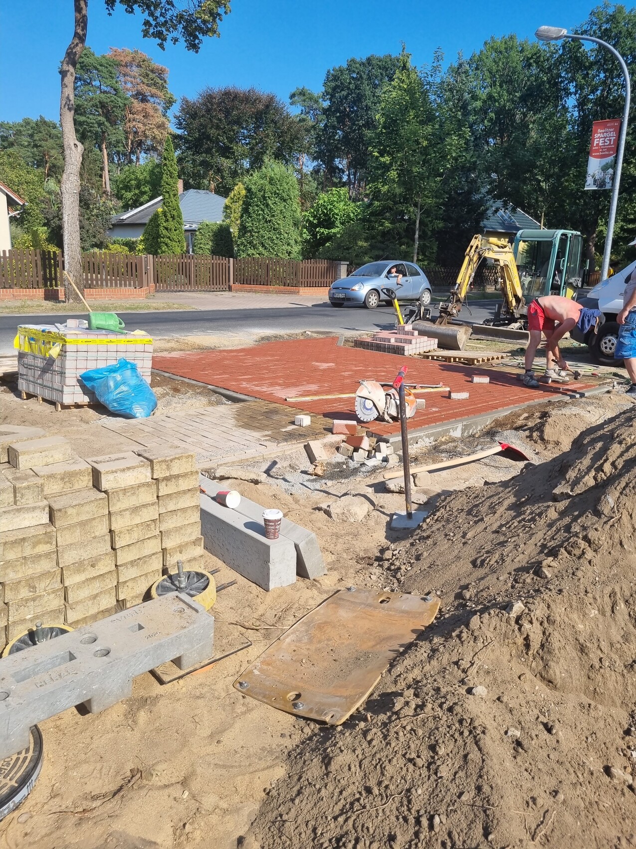 Bauarbeiter verlegen rote Pflastersteine; Paletten, Bauwerkzeuge und Straßenrandbaustelle.